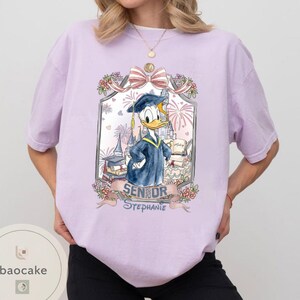May include: A light purple t-shirt featuring a graduation-themed graphic of Donald Duck in a cap and gown. The graphic includes the text "Senior Stephanie" and decorative elements like fireworks and flowers.