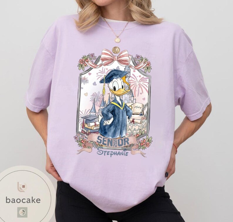 May include: A light purple t-shirt featuring a graduation-themed graphic of Donald Duck in a cap and gown. The graphic includes the text "Senior Stephanie" and decorative elements like fireworks and flowers.