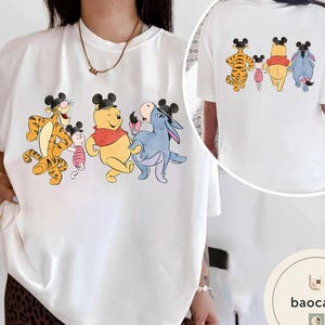 May include: White t-shirt featuring a colorful graphic of Winnie the Pooh characters: Tigger, Piglet, Winnie the Pooh, and Eeyore, all wearing Mickey Mouse ears. The back of the shirt also has the same characters. A fun, whimsical design.