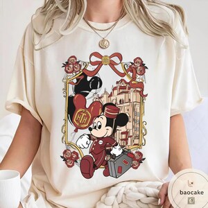 May include: Cream-colored t-shirt featuring a cartoon of Mickey Mouse in a bellhop uniform, carrying a suitcase. The design includes a framed image of a building, red roses, and balloons with the letters "HTH". A red bow is at the top of the frame.