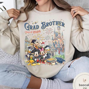 May include: Cream-coloured sweatshirt with a colourful graphic of Disney characters in graduation attire. The text "Grad Brother Class of 2026" is above an illustration of a castle and fireworks.