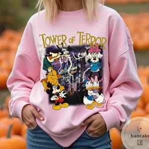 May include: Pink sweatshirt featuring a graphic of Disney characters, including Mickey Mouse, Donald Duck, and Minnie Mouse, with the text "Tower of Terror." The design includes a spooky building and lightning bolts.