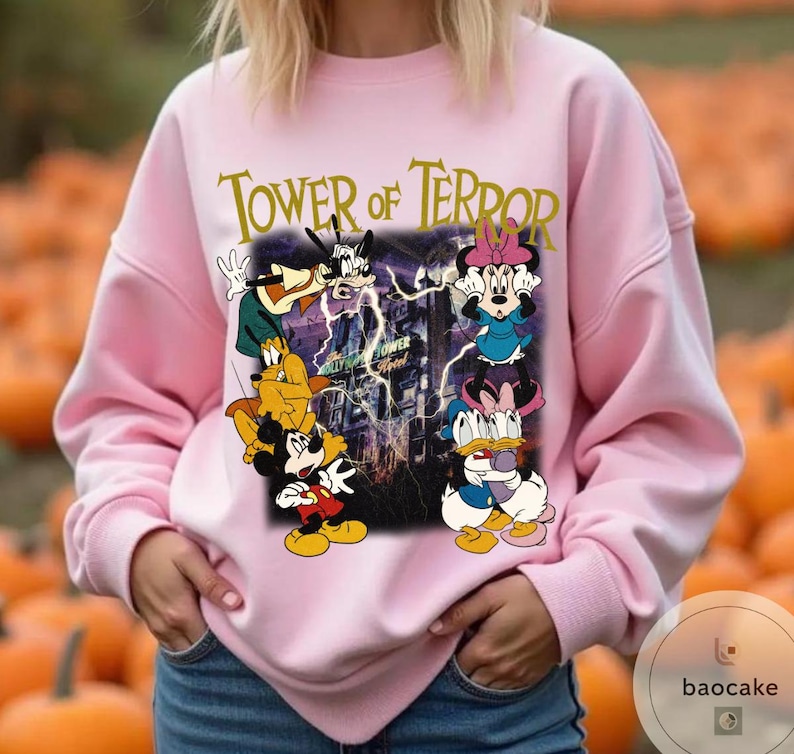 May include: Pink sweatshirt featuring a graphic of Disney characters, including Mickey Mouse, Donald Duck, and Minnie Mouse, with the text "Tower of Terror." The design includes a spooky building and lightning bolts.