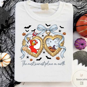 May include: White t-shirt with a Halloween graphic. The design includes a heart-shaped locket with cartoon characters, bats, spiders, and pumpkins. The text "The most scariest place on earth" is printed below the locket.