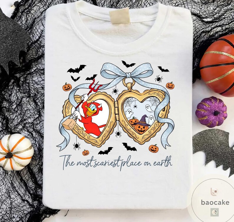 May include: White t-shirt with a Halloween graphic. The design includes a heart-shaped locket with cartoon characters, bats, spiders, and pumpkins. The text "The most scariest place on earth" is printed below the locket.