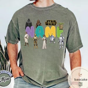 May include: A moss green t-shirt featuring Star Wars characters and the word "NAME" in colorful bubble letters. The Star Wars logo is above the name. Characters include Darth Vader, Chewbacca, Yoda, and others.