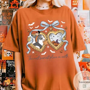 May include: Orange t-shirt with a Halloween design. The design features two heart-shaped lockets with cartoon characters, bats, spiders, and pumpkins. The text "The most scariest place on earth" is printed below the lockets.