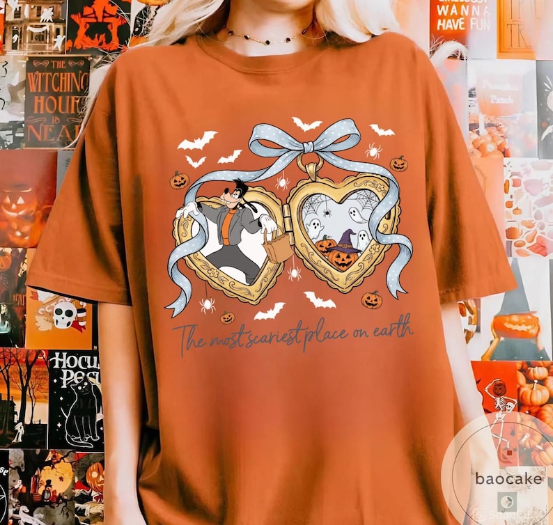 May include: Orange t-shirt with a Halloween design. The design features two heart-shaped lockets with cartoon characters, bats, spiders, and pumpkins. The text "The most scariest place on earth" is printed below the lockets.