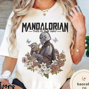 Puede incluir: Camiseta color crema con un dise&ntilde;o de The Mandalorian y Grogu, rodeados de flores y mariposas. El texto "MANDALORIAN - THIS IS THE WAY" est&aacute; impreso en negro en la parte superior. El dise&ntilde;o est&aacute; centrado en la camiseta.