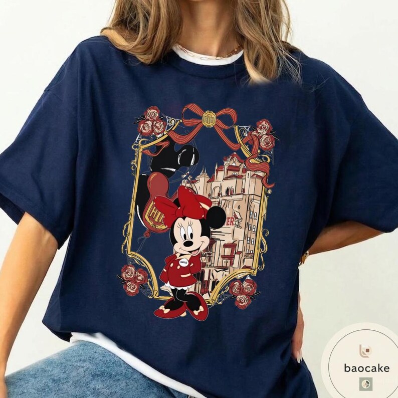 May include: Navy blue t-shirt featuring a framed graphic of Minnie Mouse in a red outfit, holding balloons, with a castle backdrop. The frame includes roses and a bow. The text "HIK" is visible on a balloon.