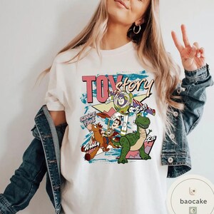 May include: Cream-colored t-shirt featuring a graphic design from the movie "Toy Story." The design includes the text "Toy Story" in pink and black, along with images of Woody, Buzz Lightyear, Rex, and other characters. The text "Since 1995" is also included.