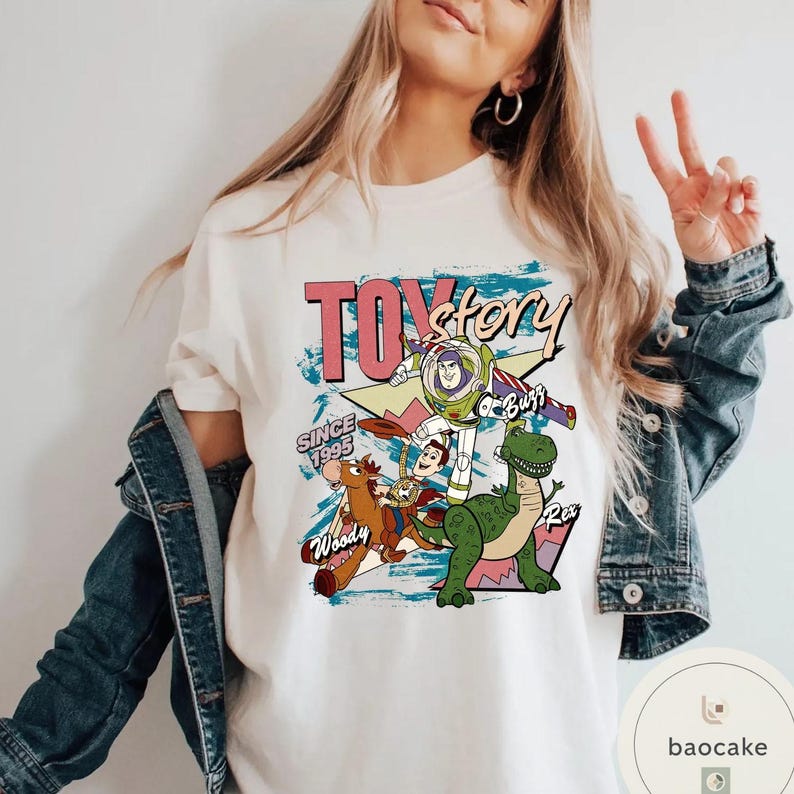 May include: Cream-colored t-shirt featuring a graphic design from the movie "Toy Story." The design includes the text "Toy Story" in pink and black, along with images of Woody, Buzz Lightyear, Rex, and other characters. The text "Since 1995" is also included.