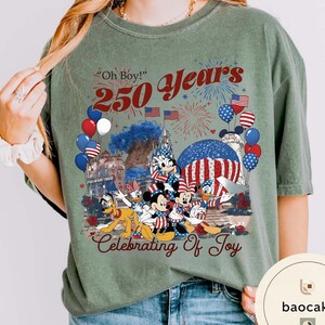 Puede incluir: Camiseta verde salvia con un gr&aacute;fico que celebra 250 a&ntilde;os. El dise&ntilde;o incluye Mickey Mouse, Minnie Mouse, Donald Duck y Pluto, junto con fuegos artificiales, globos y banderas americanas. El texto incluye "Oh Boy!" y "Celebrating of Joy."