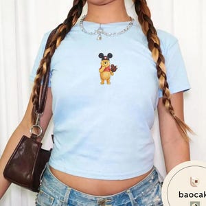 May include: A light blue short-sleeve t-shirt featuring a small graphic of a yellow bear wearing black mouse ears and holding a brown mouse-shaped treat. The shirt is paired with a silver chain necklace and a dark brown shoulder bag.
