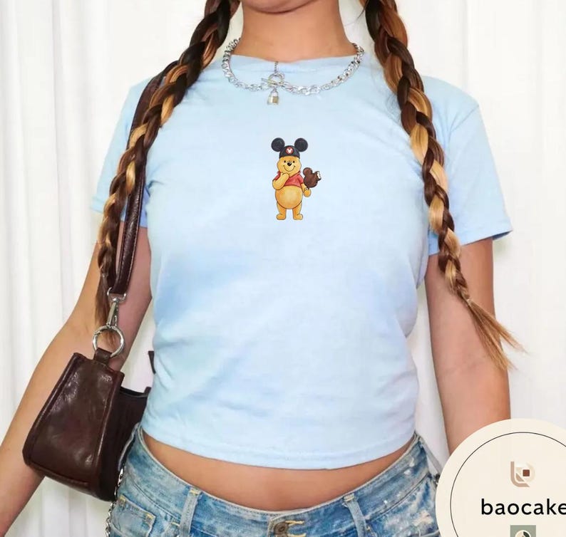 May include: A light blue short-sleeve t-shirt featuring a small graphic of a yellow bear wearing black mouse ears and holding a brown mouse-shaped treat. The shirt is paired with a silver chain necklace and a dark brown shoulder bag.