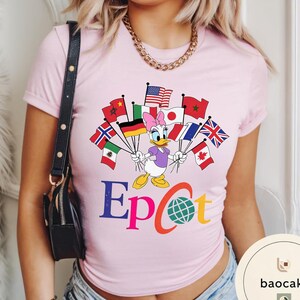 May include: A pale pink t-shirt with a Daisy Duck graphic holding flags from different nations. The word "Epcot" is printed in colourful letters below the character. A gold chain and black purse are also visible.