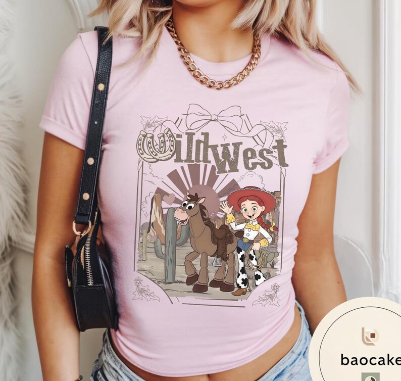 Pu&ograve; includere: T-shirt rosa chiaro con un'illustrazione a cartoni animati raffigurante una cowgirl, un cavallo e un paesaggio desertico, con la scritta "Wild West". La maglietta ha maniche corte e girocollo.