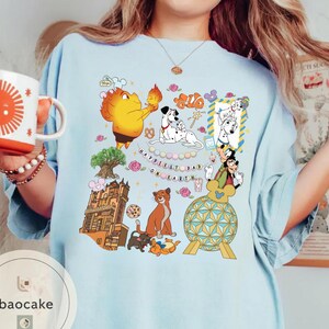 May include: Light blue t-shirt featuring a colorful graphic with cartoon characters, including Dalmatian dogs, Mickey Mouse, and a cat. The text "Happiest Dad on Earth" is also visible. A white mug with a sun design is held in the image.