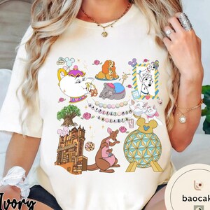 May include: Ivory-colored t-shirt featuring colorful cartoon illustrations of Disney characters and the text "Happiest Mom on Earth." The design includes a teapot, dogs, cats, a kangaroo, and a castle.