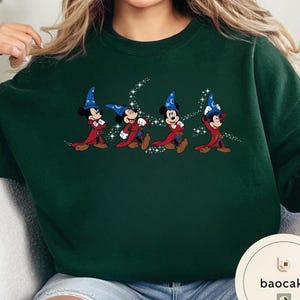May include: Dark green sweatshirt featuring a graphic of Mickey Mouse in a wizard costume, with a blue hat and red robe. The design includes a starry trail. The sweatshirt is a solid color.