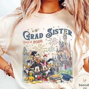 May include: Cream-coloured t-shirt with a colourful graphic. The design features the text "Grad Sister Class of 2026" and Disney characters in graduation attire, a castle, fireworks, and books. The graphic uses shades of blue, red, yellow, and black.