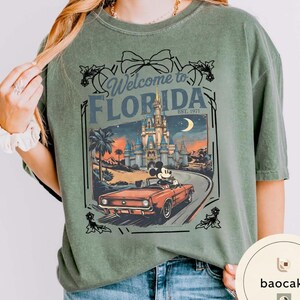 May include: Sage green t-shirt with a graphic featuring a vintage car driven by Mickey Mouse on a road leading to a castle. The text reads "Welcome to Florida" and "Est. 1971". The design is framed with decorative elements.