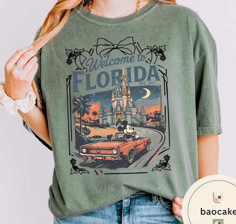 May include: Sage green t-shirt with a graphic featuring a vintage car driven by Mickey Mouse on a road leading to a castle. The text reads "Welcome to Florida" and "Est. 1971". The design is framed with decorative elements.