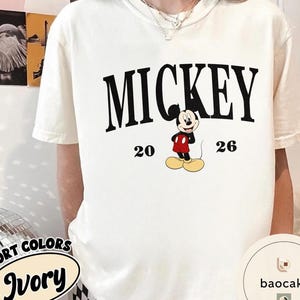 May include: Ivory-colored t-shirt featuring the word "MICKEY" in bold black letters, with the numbers "20" and "26" on either side of a cartoon Mickey Mouse graphic. The shirt has short sleeves.