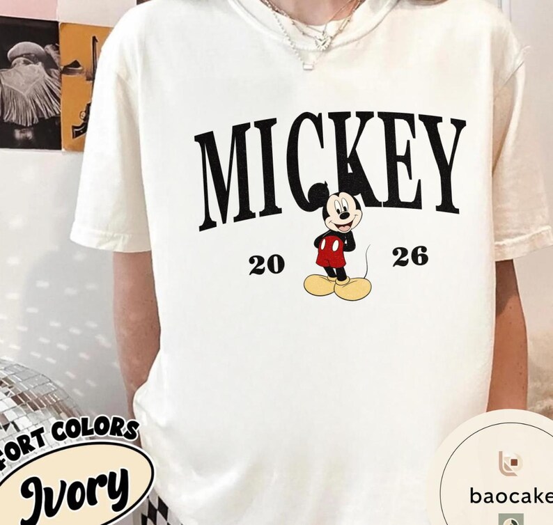 May include: Ivory-colored t-shirt featuring the word "MICKEY" in bold black letters, with the numbers "20" and "26" on either side of a cartoon Mickey Mouse graphic. The shirt has short sleeves.