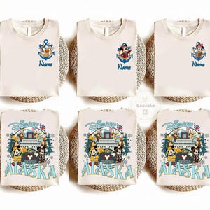 May include: Cream-colored t-shirts with Disney character designs. Some shirts feature an anchor with a character and the word "Name" in blue. Other shirts have a Disney Alaska 2024 design with Mickey and friends.