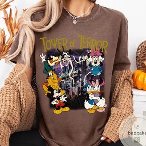 May include: Brown t-shirt featuring a graphic of Disney characters, including Mickey Mouse, Goofy, and Minnie Mouse, with the text "Tower of Terror." The design includes lightning and the Hollywood Tower Hotel.