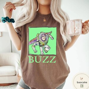 K&ouml;nnte beinhalten: Ein braunes Kurzarm-T-Shirt mit einer Grafik der Figur Buzz Lightyear in einem hellgr&uuml;nen Quadrat. Die Figur tr&auml;gt einen wei&szlig;en, gr&uuml;nen und violetten Raumanzug mit Fl&uuml;geln. Das Wort BUZZ ist darunter in gro&szlig;en gr&uuml;nen Buchstaben aufgedruckt.