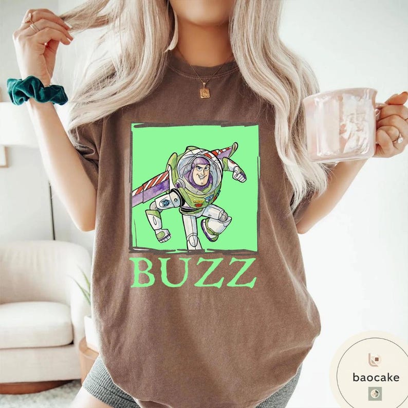 K&ouml;nnte beinhalten: Ein braunes Kurzarm-T-Shirt mit einer Grafik der Figur Buzz Lightyear in einem hellgr&uuml;nen Quadrat. Die Figur tr&auml;gt einen wei&szlig;en, gr&uuml;nen und violetten Raumanzug mit Fl&uuml;geln. Das Wort BUZZ ist darunter in gro&szlig;en gr&uuml;nen Buchstaben aufgedruckt.