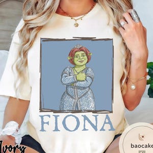 May include: Ivory-colored t-shirt featuring a graphic of Princess Fiona from Shrek. The illustration shows Fiona with red hair, a tiara, and a blue dress, set against a blue background. The name "FIONA" is printed in blue below the image.