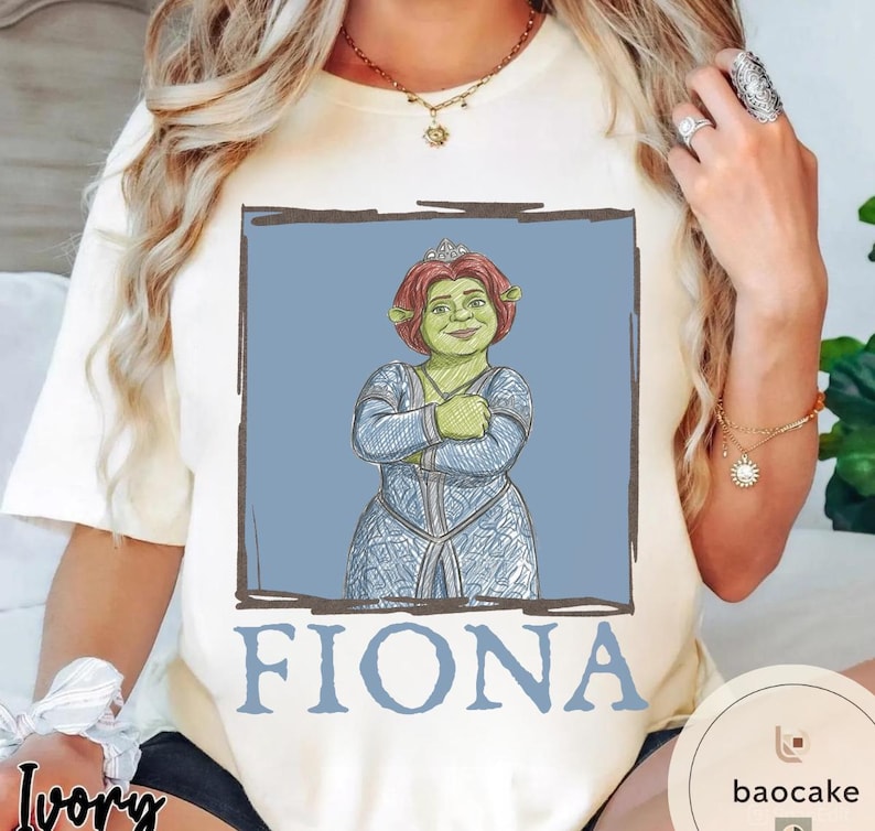 May include: Ivory-colored t-shirt featuring a graphic of Princess Fiona from Shrek. The illustration shows Fiona with red hair, a tiara, and a blue dress, set against a blue background. The name "FIONA" is printed in blue below the image.