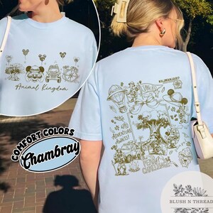 May include: Light blue Chambray t-shirt with a gold graphic design featuring the text "Animal Kingdom" on the front. The back of the shirt has a detailed illustration with the text "Adventure Awaits!" and "Peak Challenge."