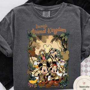 May include: Dark gray t-shirt featuring a graphic of Disney's Animal Kingdom characters, including Mickey and Minnie Mouse, Donald Duck, Goofy, and Pluto, in safari attire. The text "Disney's Animal Kingdom" is displayed above the characters.