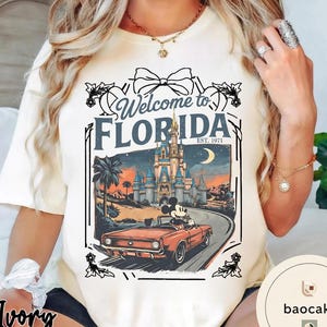 May include: Ivory-colored t-shirt featuring a graphic of Mickey Mouse driving a red convertible towards a castle at sunset. The text "Welcome to Florida" and "EST. 1971" are also displayed.