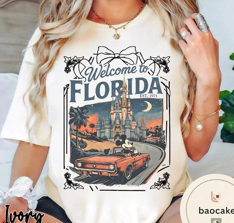 May include: Ivory-colored t-shirt featuring a graphic of Mickey Mouse driving a red convertible towards a castle at sunset. The text "Welcome to Florida" and "EST. 1971" are also displayed.