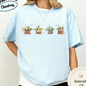 May include: Light blue t-shirt featuring four cartoon characters in different outfits. The characters are green with large eyes and are surrounded by small stars. The shirt has the word "Chambray" printed on it.