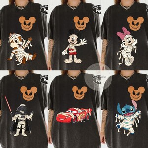 May include: A collection of black graphic t-shirts featuring various cartoon characters in Halloween mummy costumes. Designs include Chip, Mickey Mouse, Minnie Mouse, Darth Vader, Lightning McQueen, and Stitch, each paired with a jack-o'-lantern Mickey Mouse head.