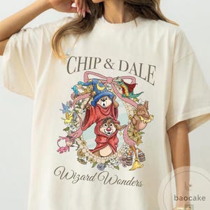 May include: Cream-colored t-shirt featuring Chip and Dale in wizard costumes, surrounded by a floral wreath and a pink ribbon. The text "Chip & Dale" and "Wizard Wonders" are printed on the shirt.