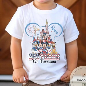 Peut inclure: T-shirt blanc avec un motif color&eacute; de personnages Disney en tenues patriotiques, dont Mickey Mouse et Donald Duck, devant un ch&acirc;teau. Le texte indique "250 Years of Freedom" et "Name".
