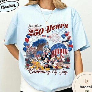 Puede incluir: Camiseta azul claro con un dise&ntilde;o que celebra 250 a&ntilde;os. El dise&ntilde;o incluye a Mickey, Minnie, Donald y Pluto con atuendos patri&oacute;ticos, junto con fuegos artificiales, globos y banderas americanas. El texto incluye "Oh Boy!" y "Celebrating Of Joy."