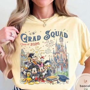 May include: A pale yellow t-shirt with a colourful graphic featuring Disney characters in graduation attire. The text "Grad Squad Class of 2026" is displayed above the characters and a castle.