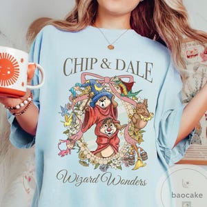 May include: Light blue t-shirt featuring Chip and Dale in wizard attire, surrounded by a floral wreath and the text "Chip & Dale" and "Wizard Wonders". The design includes a wizard hat, wand, and other whimsical elements.