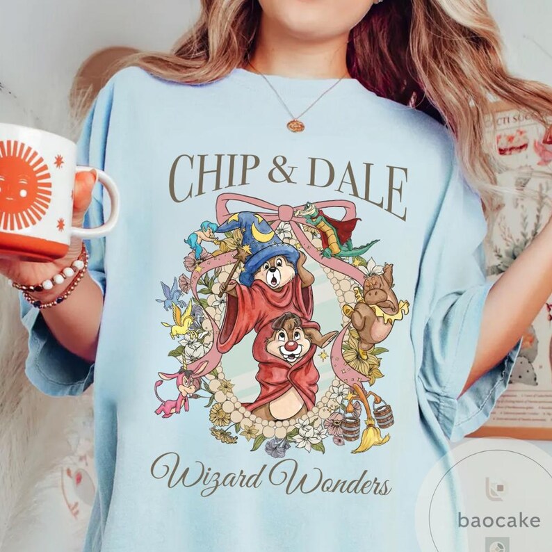 May include: Light blue t-shirt featuring Chip and Dale in wizard attire, surrounded by a floral wreath and the text "Chip & Dale" and "Wizard Wonders". The design includes a wizard hat, wand, and other whimsical elements.
