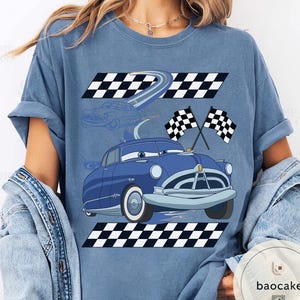 May include: A blue t-shirt featuring a cartoon car design with racing flags and checkered patterns. The car is blue with white accents, and the design includes a racing track and checkered flags. The shirt is a light blue color.