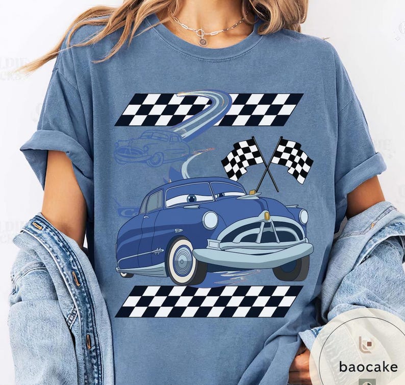May include: A blue t-shirt featuring a cartoon car design with racing flags and checkered patterns. The car is blue with white accents, and the design includes a racing track and checkered flags. The shirt is a light blue color.