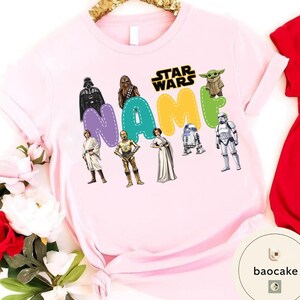 May include: A light pink t-shirt featuring Star Wars characters and the word "NAME" in colorful letters. Characters include Darth Vader, Chewbacca, Yoda, and a Stormtrooper. The Star Wars logo is also visible.